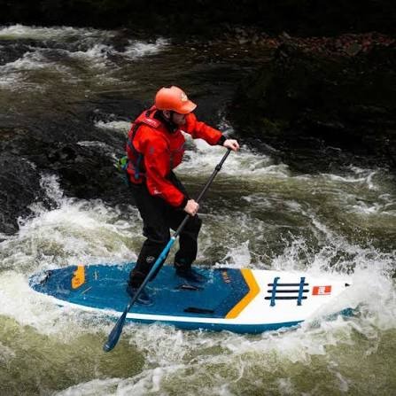 THe rigors of whitewater paddling with man on rapids standing on paddleboard with orange safety gear.