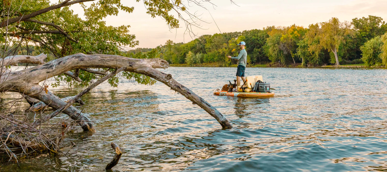 A man fishing a river on GO boat fish with fallen trees near and trees on opposite bank.