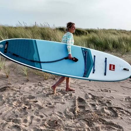 A woman carrying her red inflatable paddleboard on the beach.