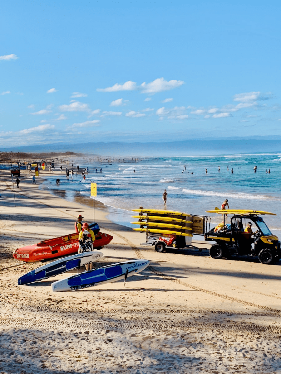 Beach scene with clear blue sky, lifeguards next to jet skis and a quad bike with surfboards. People enjoy swimming in the gentle ocean waves.